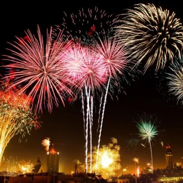 Colorful fireworks bursting over a city skyline at night, with red, pink, and gold trails lighting the sky.