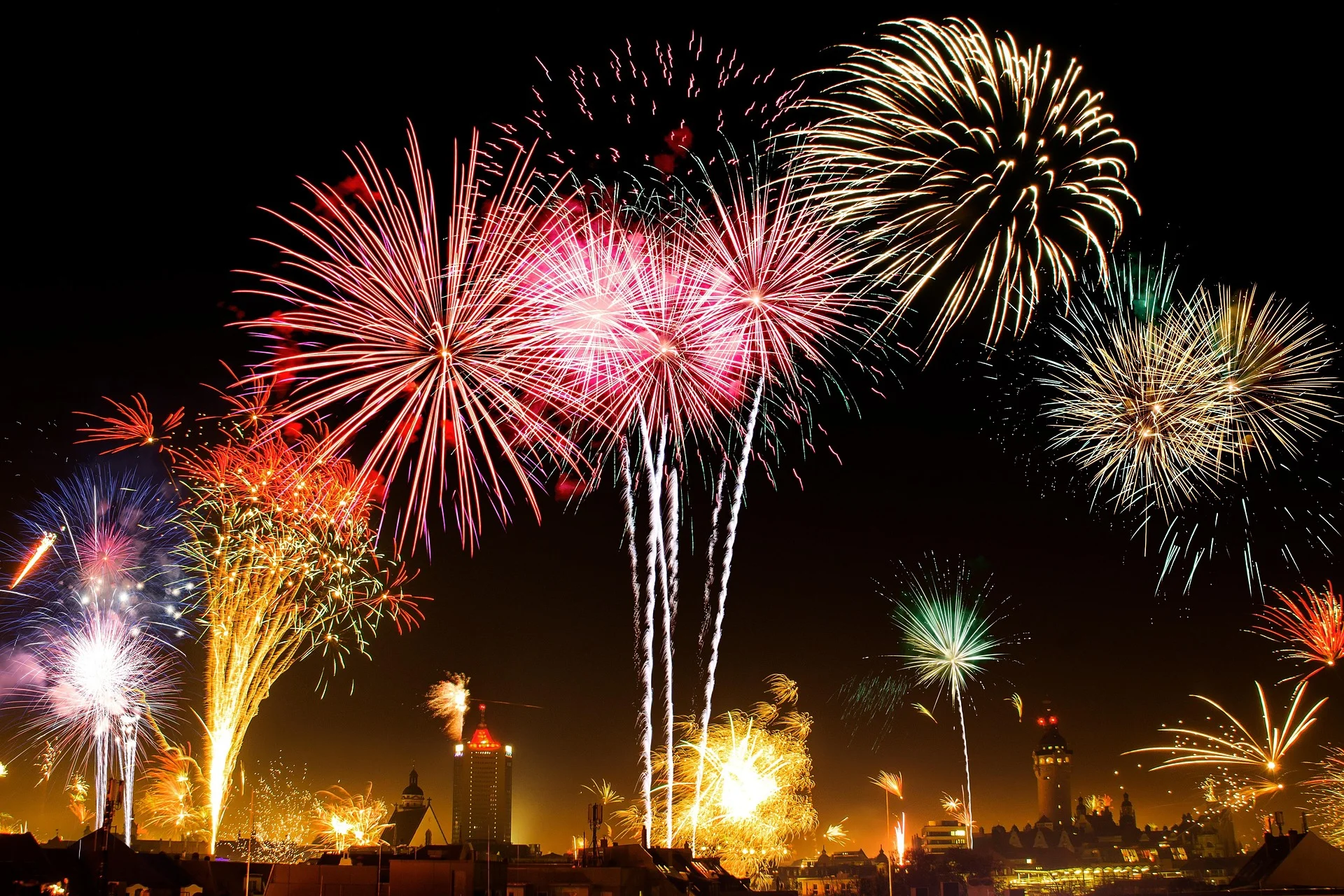 Colorful fireworks bursting over a city skyline at night, with red, pink, and gold trails lighting the sky.