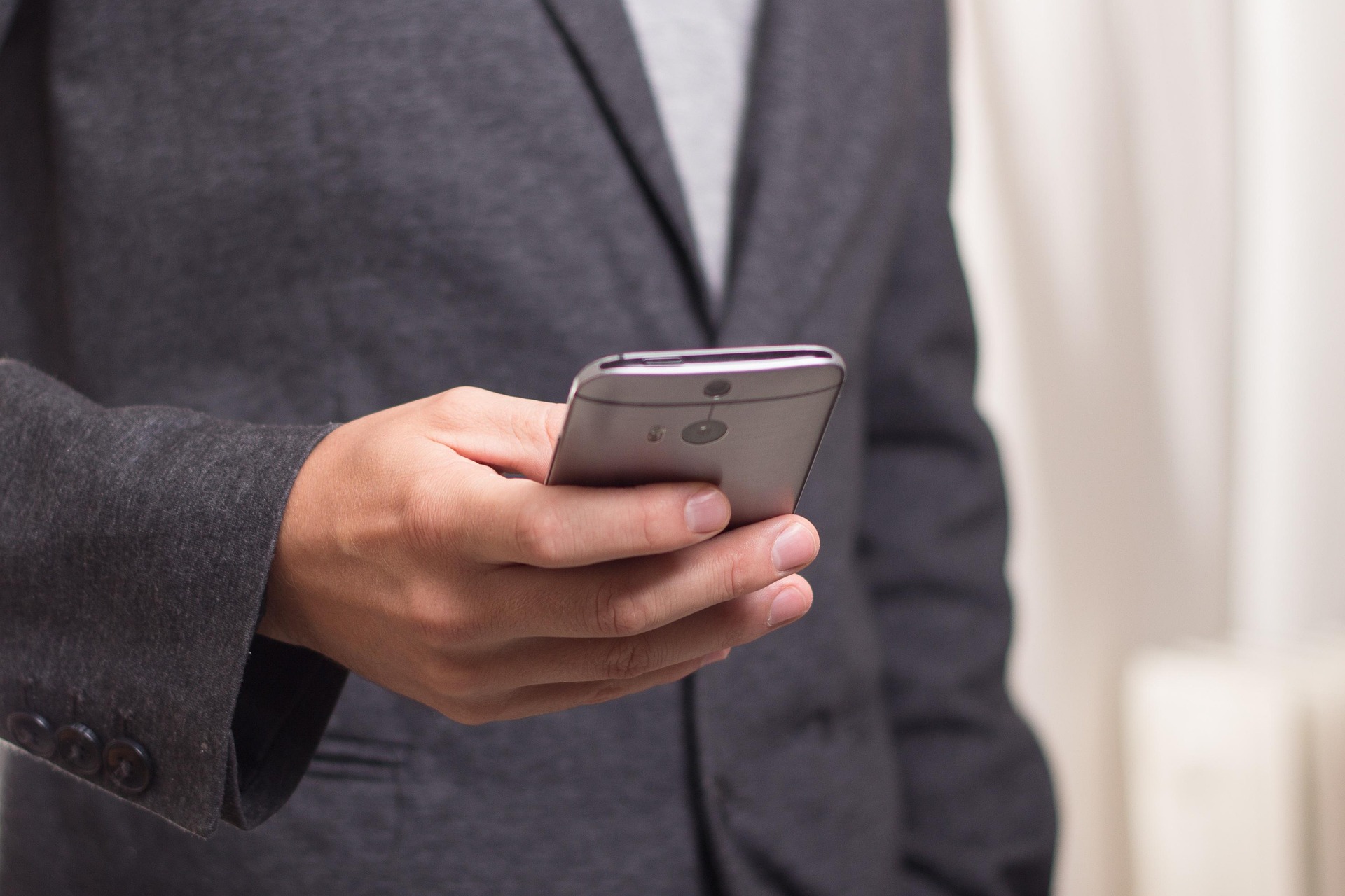 Hands holding a silver smartphone in a dark blazer; close-up of device and hands.