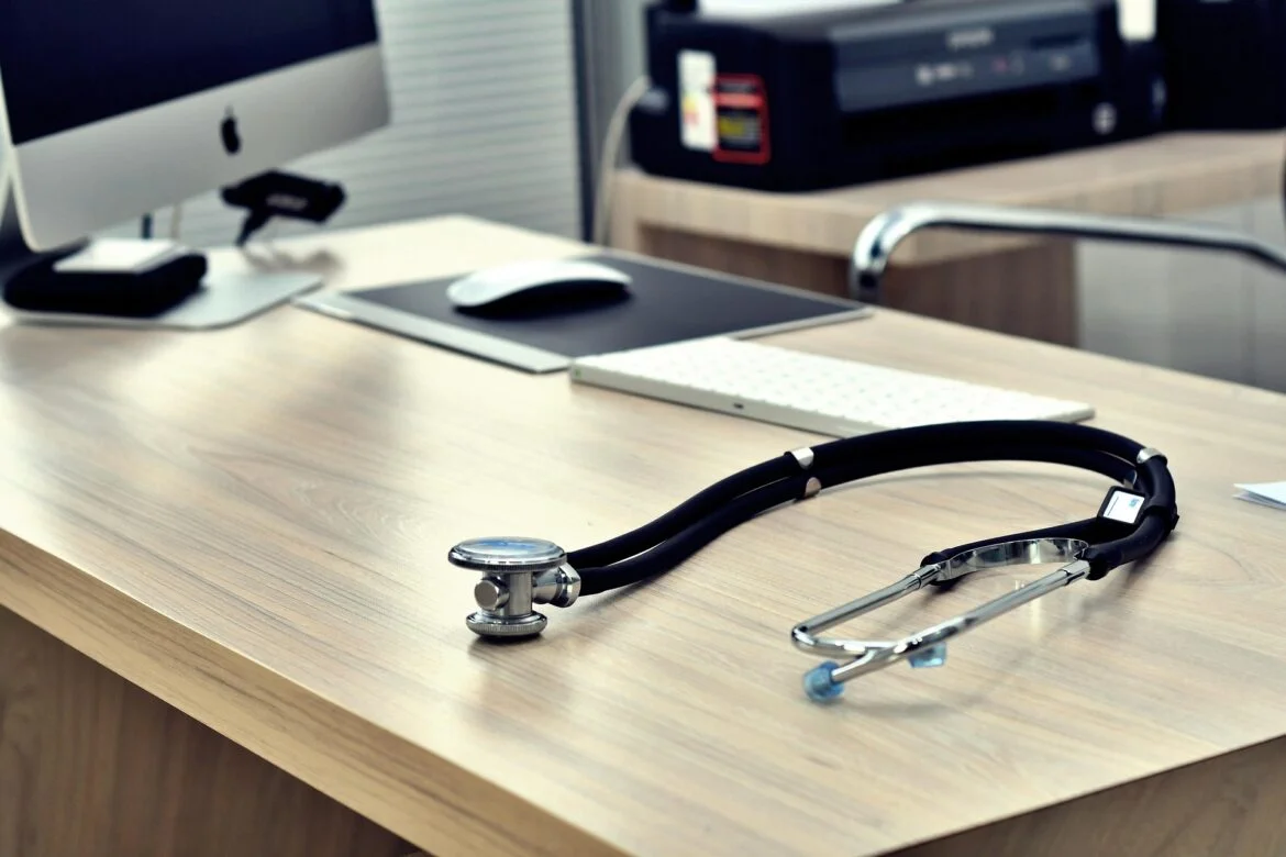 Stethoscope on a wooden desk in a medical office, with keyboard, mouse, monitor, and printer in the background.