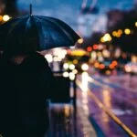Person in a dark coat holding a blue umbrella on a rain-soaked city street at night with blurred colorful lights in the background