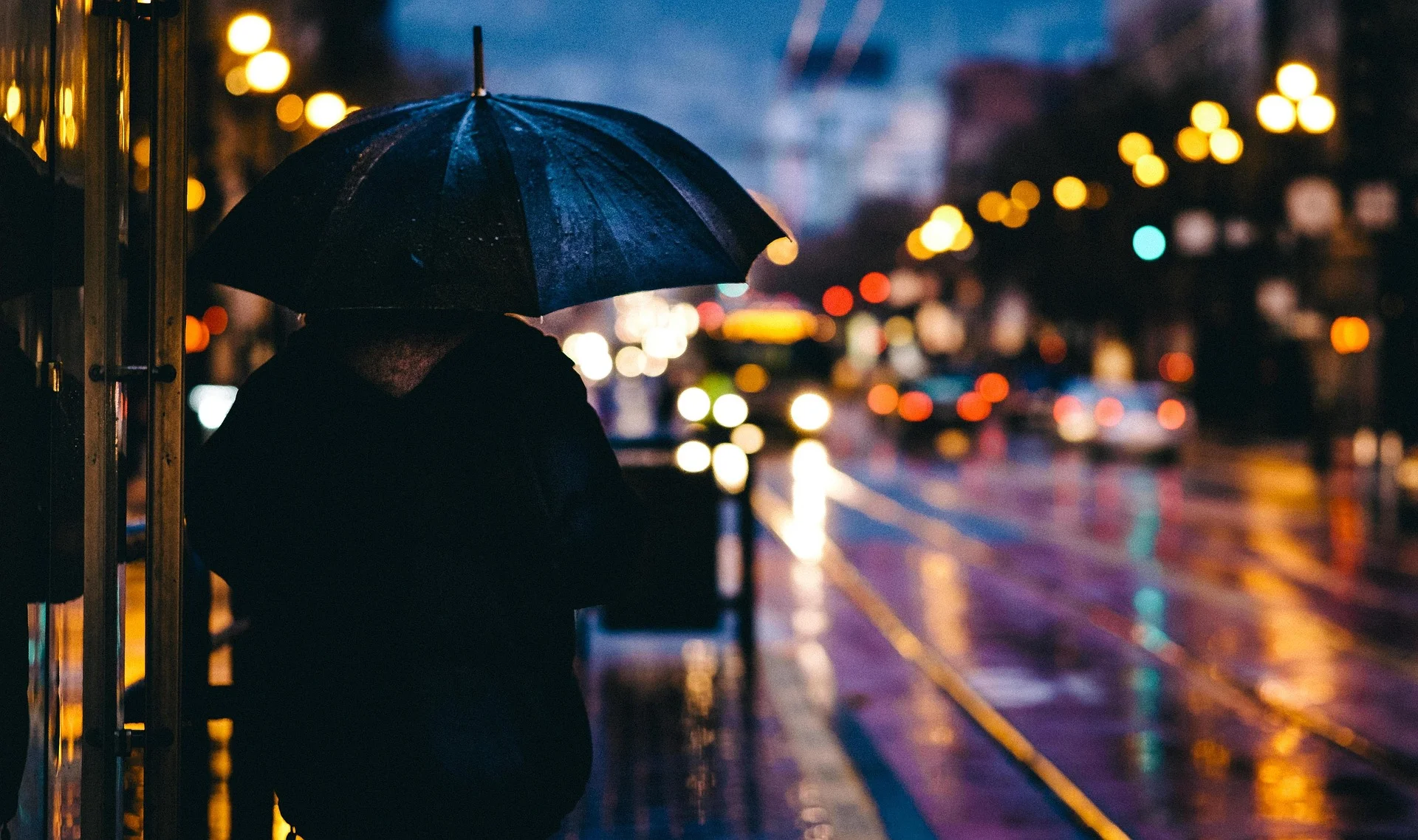Person in a dark coat holding a blue umbrella on a rain-soaked city street at night with blurred colorful lights in the background
