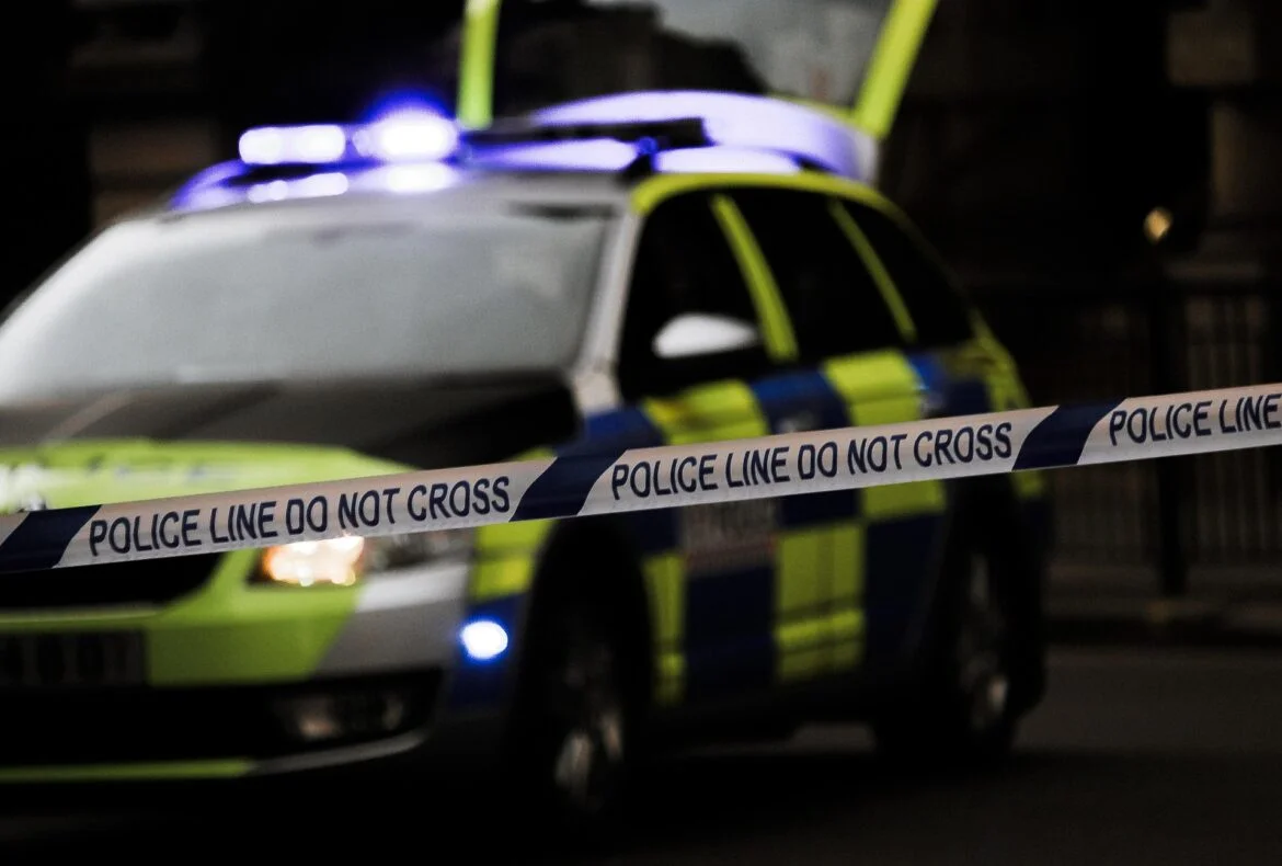 Police car with blue lights behind a 'Police Line Do Not Cross' barrier tape at a nighttime scene