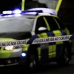 Police car with blue lights behind a 'Police Line Do Not Cross' barrier tape at a nighttime scene