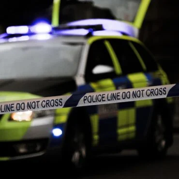 Police car with blue lights behind a 'Police Line Do Not Cross' barrier tape at a nighttime scene