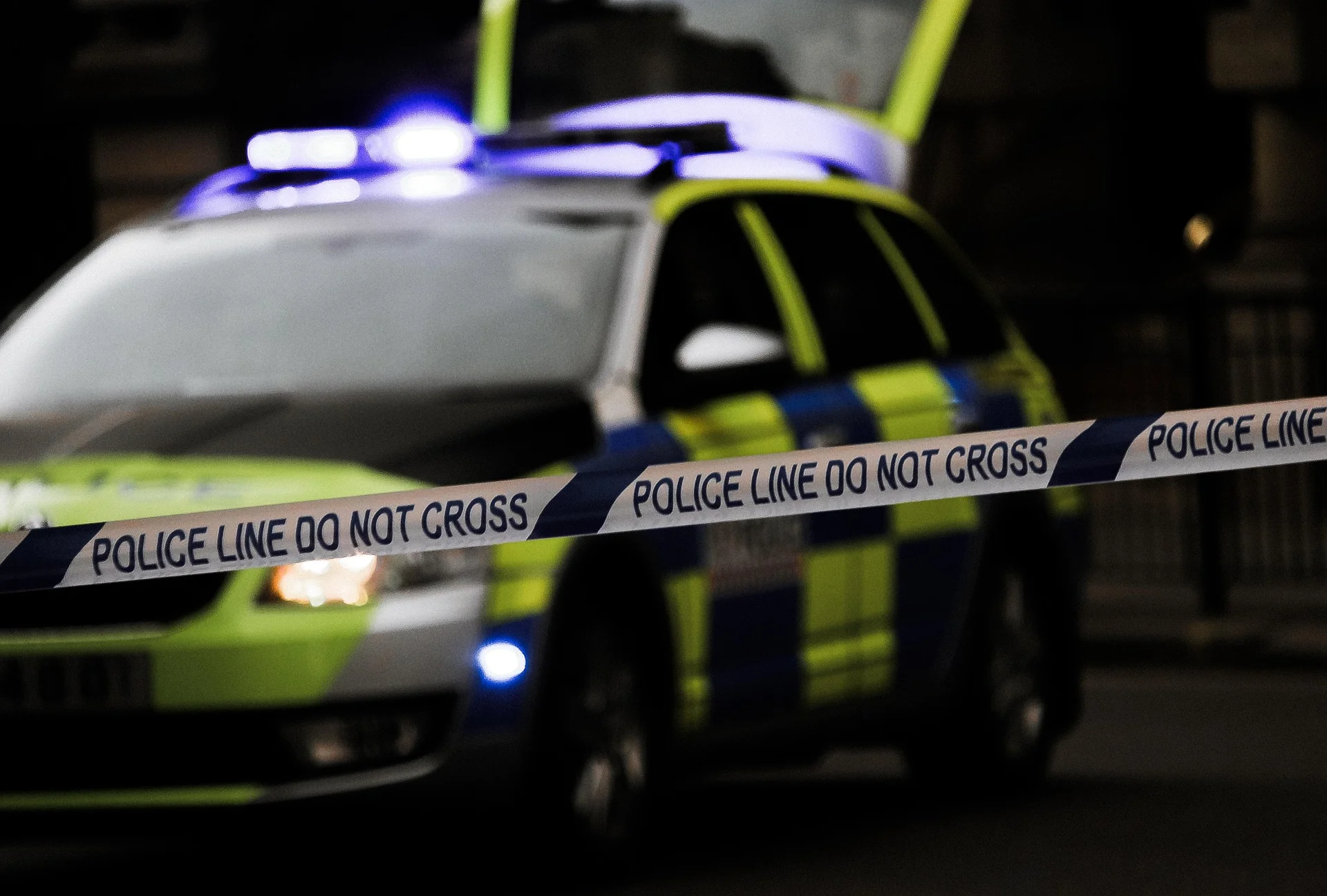 Police car with blue lights behind a 'Police Line Do Not Cross' barrier tape at a nighttime scene