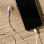 Smartphone with black screen in a clear case on a wooden table, with white wired earphones around it.