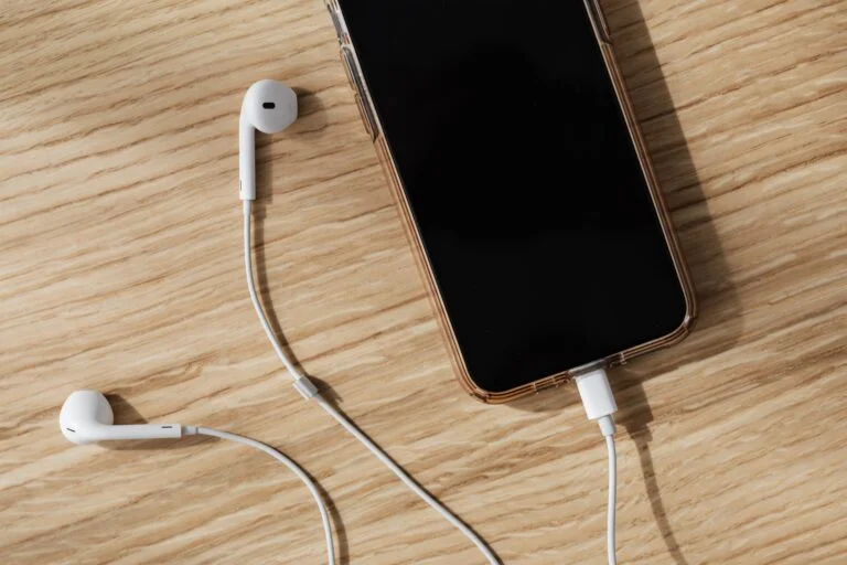 Smartphone with black screen in a clear case on a wooden table, with white wired earphones around it.