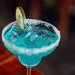 Turquoise margarita in a salted rim glass with a lime wheel garnish and ice-filled drink close up.
