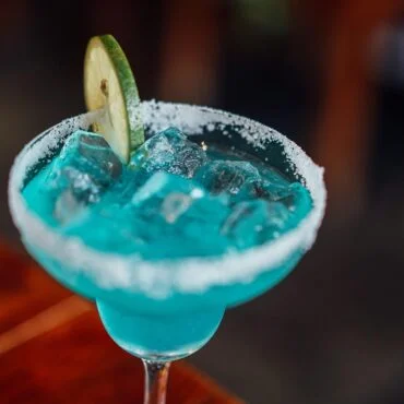 Turquoise margarita in a salted rim glass with a lime wheel garnish and ice-filled drink close up.