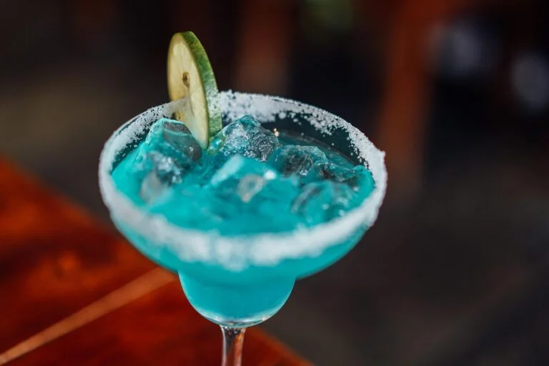 Turquoise margarita in a salted rim glass with a lime wheel garnish and ice-filled drink close up.