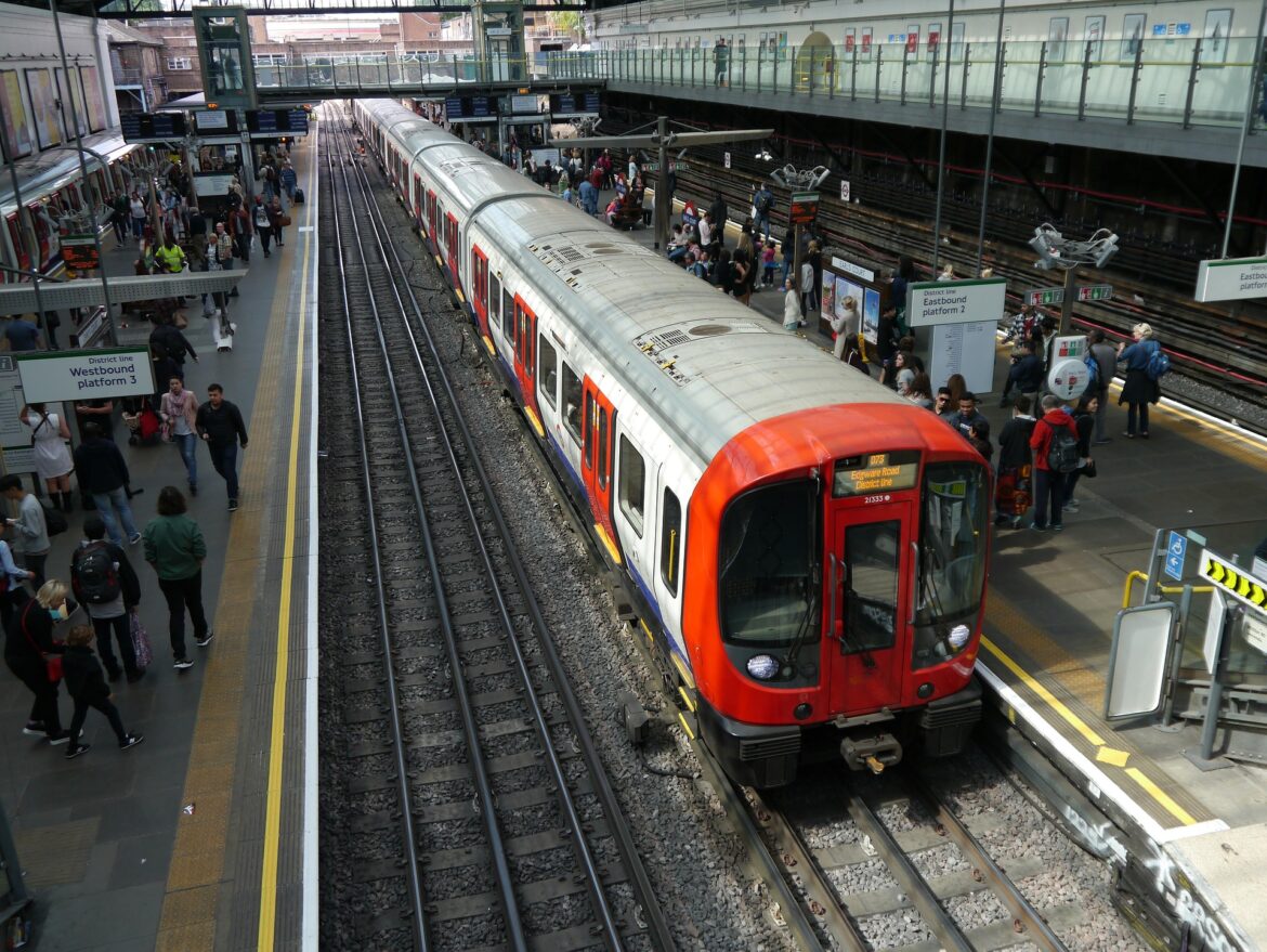 Crowded train platform with a red-fronted train at the edge and many commuters waiting or walking along the platform.