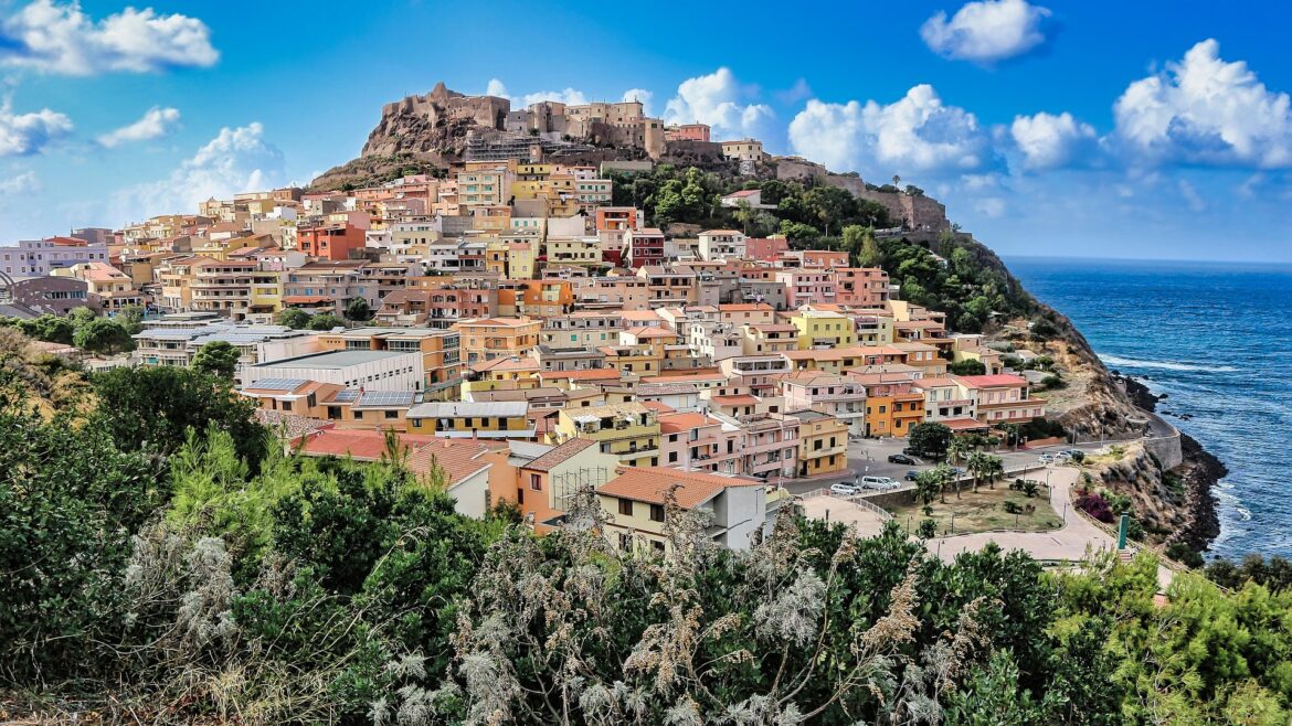 Colorful hillside town built into a cliff by the blue sea, with a fortress on top under a bright sky.
