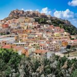 Colorful hillside town built into a cliff by the blue sea, with a fortress on top under a bright sky.