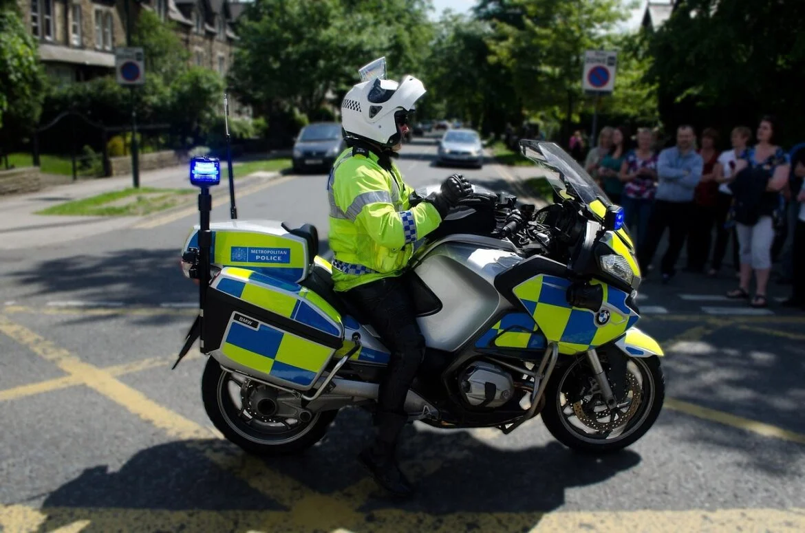 Police motorcycle with blue lights on, officer in high-visibility jacket seated on a BMW bike beside a crowd on a city street.