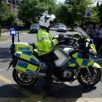 Police motorcycle with blue lights on, officer in high-visibility jacket seated on a BMW bike beside a crowd on a city street.