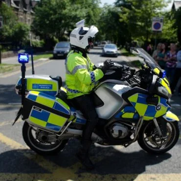 Police motorcycle with blue lights on, officer in high-visibility jacket seated on a BMW bike beside a crowd on a city street.