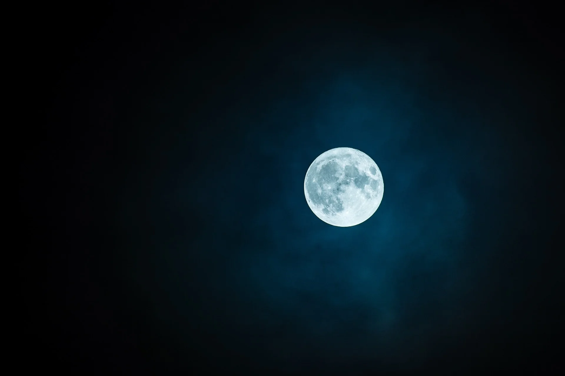 Full moon glowing blue in a dark night sky with wispy clouds.