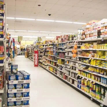 A supermarket aisle filled with shelves of canned goods, primarily Goya, on both sides under bright overhead lights.