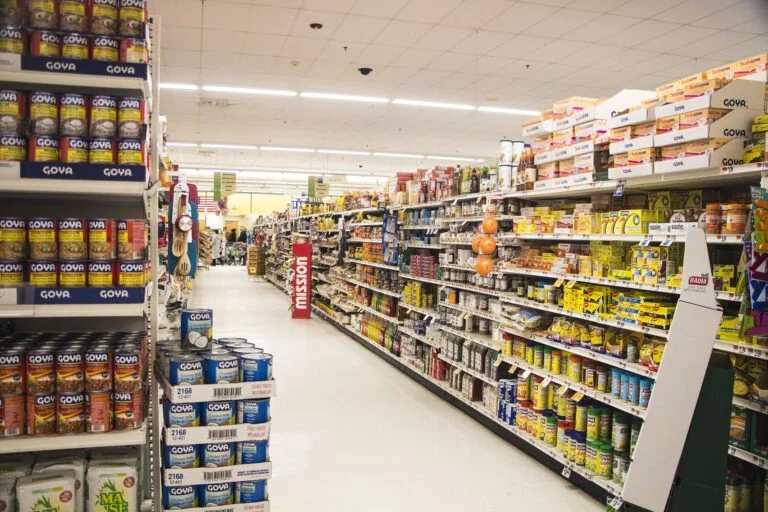 A supermarket aisle filled with shelves of canned goods, primarily Goya, on both sides under bright overhead lights.