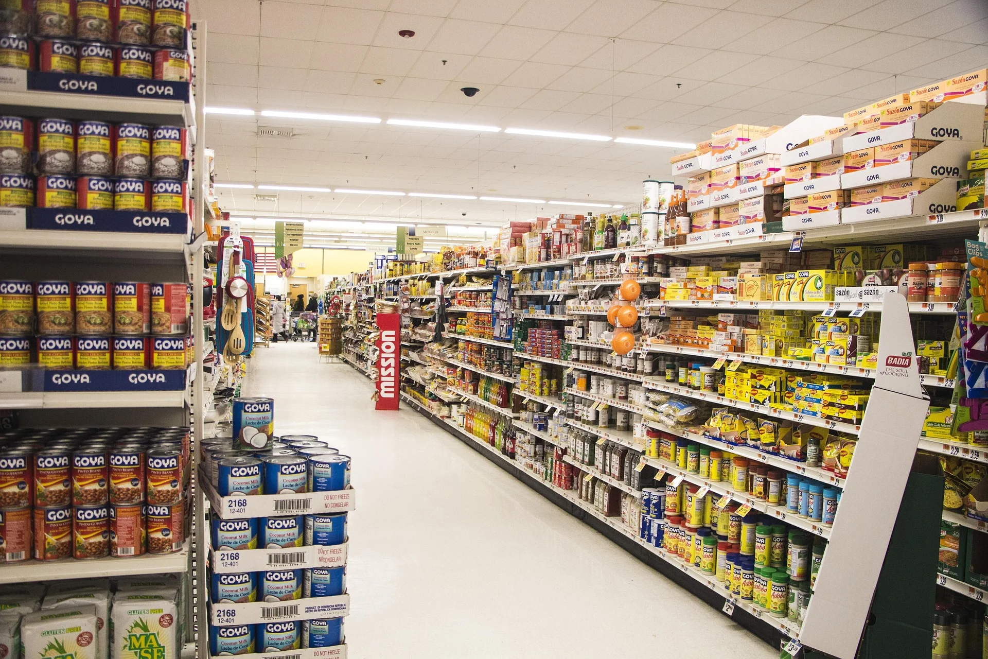A supermarket aisle filled with shelves of canned goods, primarily Goya, on both sides under bright overhead lights.