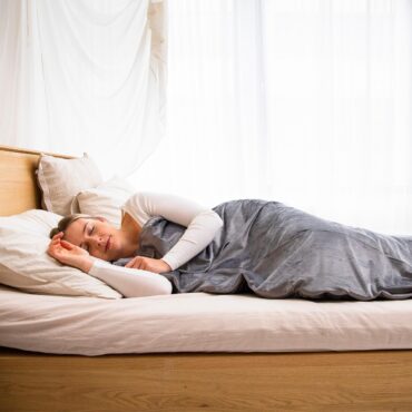 Woman sleeping on a white bed in a bright, airy bedroom with sheer curtains and gray bedding
