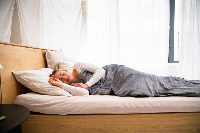 Woman sleeping on a white bed in a bright, airy bedroom with sheer curtains and gray bedding