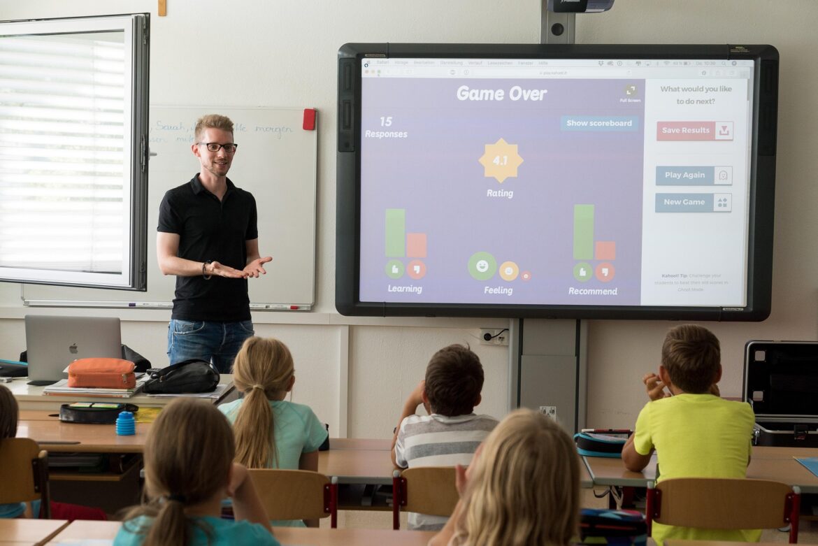 Teacher presenting in a classroom beside a large screen showing a 'Game Over' results screen with score and options at right edge.