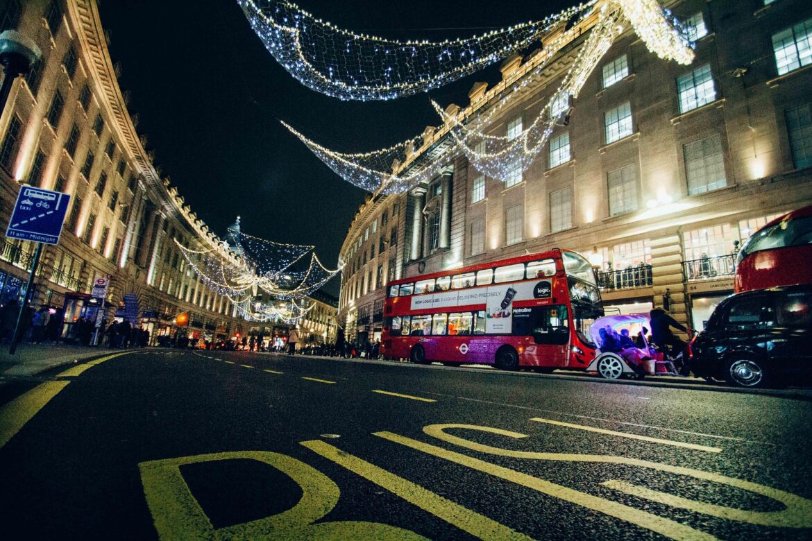 Nighttime city street with decorative string lights, a red double-decker bus, and pedestrians.