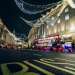 Nighttime city street with decorative string lights, a red double-decker bus, and pedestrians.