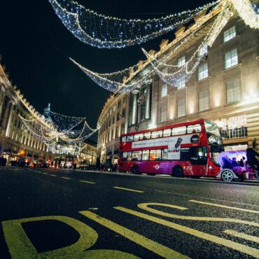 Nighttime city street with decorative string lights, a red double-decker bus, and pedestrians.