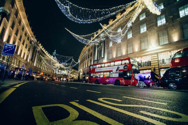 Nighttime city street with decorative string lights, a red double-decker bus, and pedestrians.