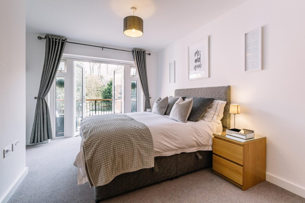 Bright modern bedroom with a gray upholstered headboard, matching bedding, and a wooden nightstand beside a lamp; glass doors open to a balcony with trees outside.