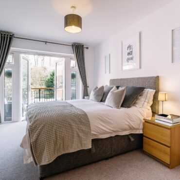 Bright modern bedroom with a gray upholstered headboard, matching bedding, and a wooden nightstand beside a lamp; glass doors open to a balcony with trees outside.