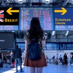 Traveler with backpack stands in an airport terminal, facing large Brexit and European Union signs above a flight information display.