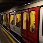 London Underground train with red doors at a dimly lit platform, passengers inside and a few people waiting on the edge.