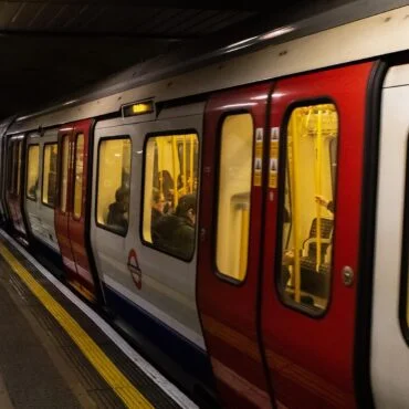 London Underground train with red doors at a dimly lit platform, passengers inside and a few people waiting on the edge.