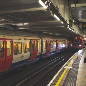 London Underground platform with a red-and-white train at the edge and passengers waiting on the platform toward the right side of the image.