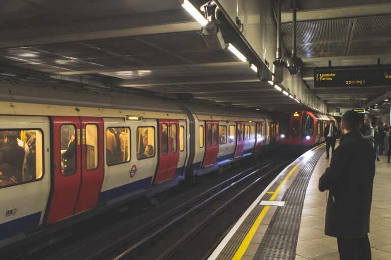 London Underground platform with a red-and-white train at the edge and passengers waiting on the platform toward the right side of the image.