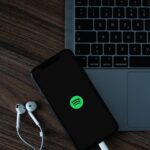 Laptop keyboard on a wooden desk with a smartphone showing the Spotify logo and white wired earbuds nearby.