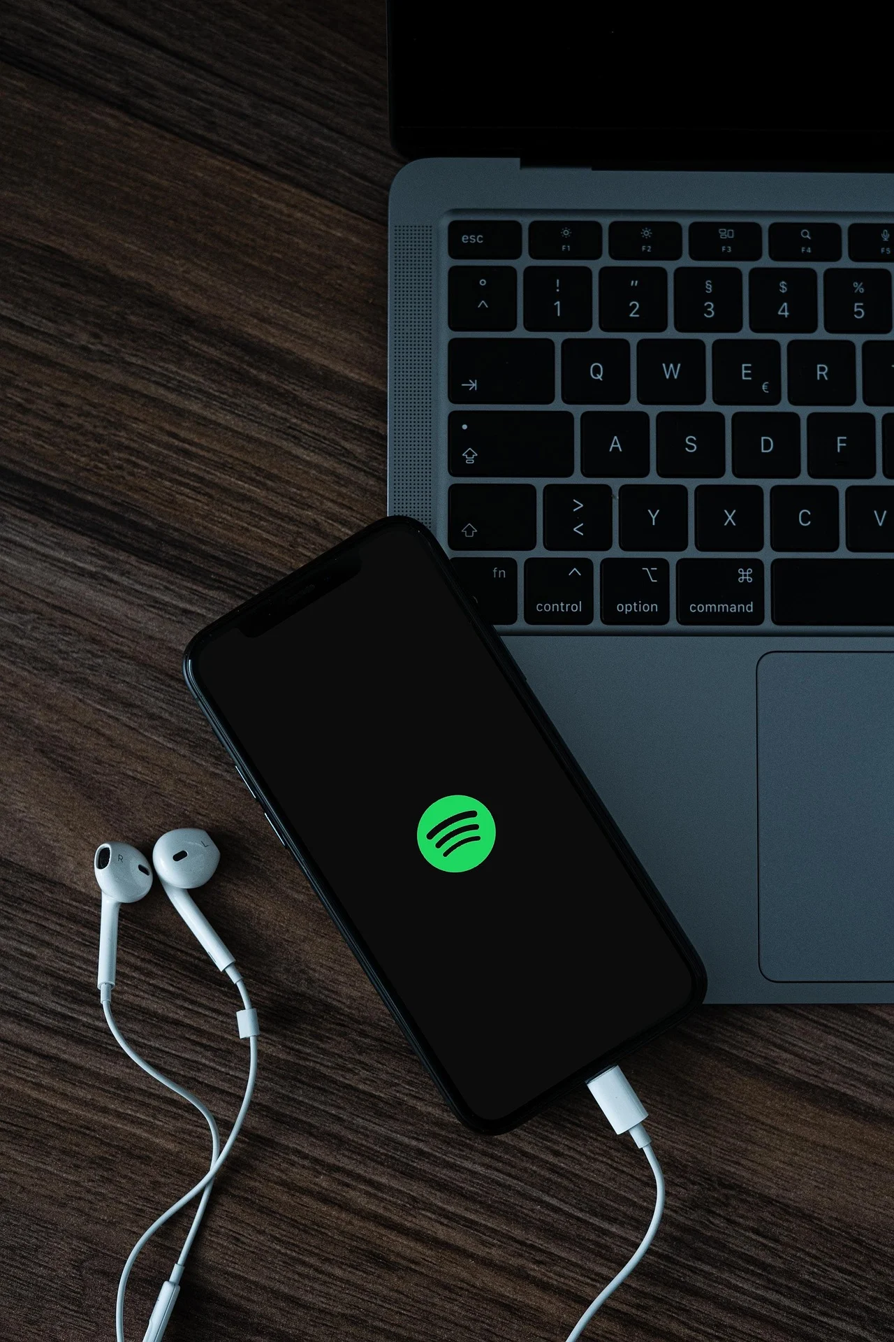 Laptop keyboard on a wooden desk with a smartphone showing the Spotify logo and white wired earbuds nearby.