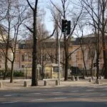 Green traffic light at a tree-lined city street with a pale yellow historic building behind a black fence.