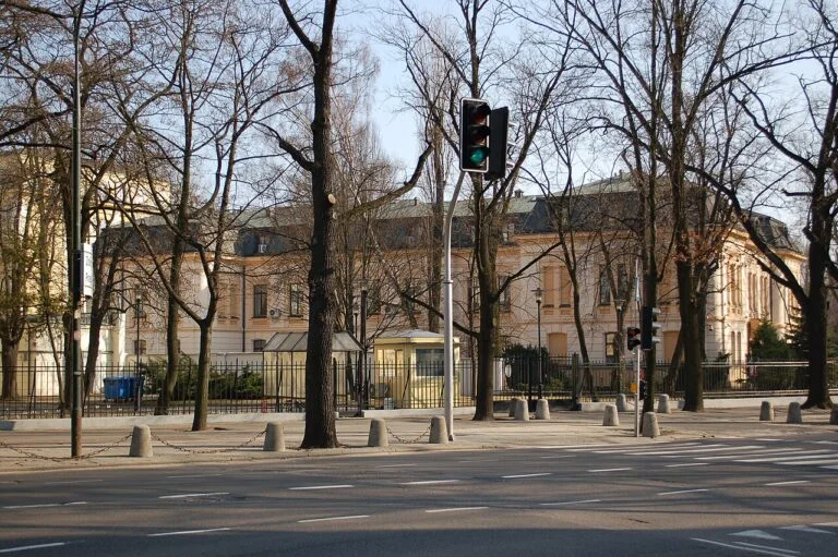 Green traffic light at a tree-lined city street with a pale yellow historic building behind a black fence.