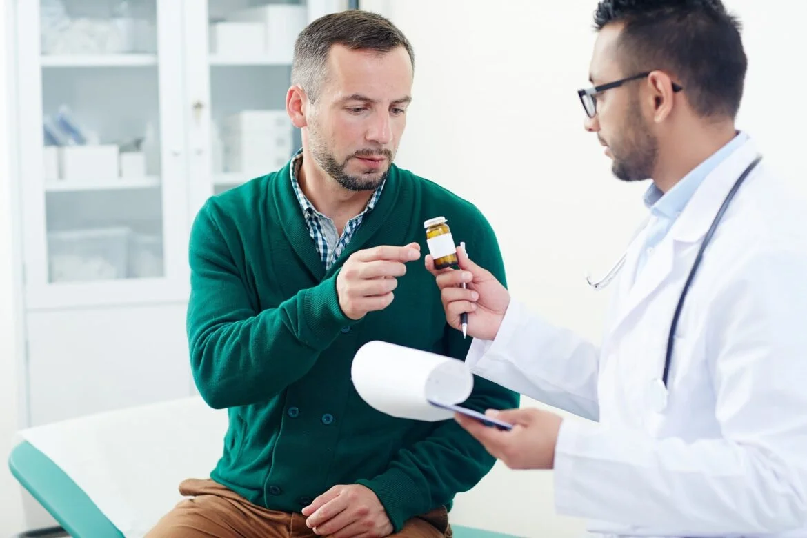 Doctor in a white coat showing a small medicine bottle to a man in a green sweater during a medical consultation.