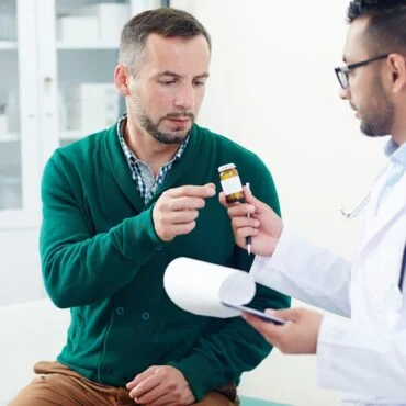 Doctor in a white coat showing a small medicine bottle to a man in a green sweater during a medical consultation.