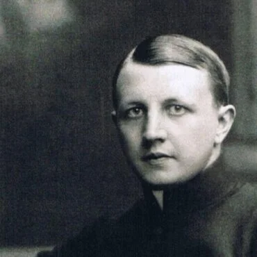 Black-and-white portrait of a person with slicked-back hair, wearing a high-collar dark garment and looking at the camera.