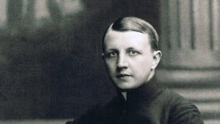 Black-and-white portrait of a person with slicked-back hair, wearing a high-collar dark garment and looking at the camera.