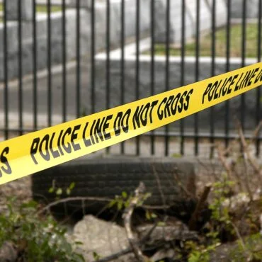 Yellow police line tape stretching diagonally across a scene with a metal fence and rubble behind it, indicating a restricted area.