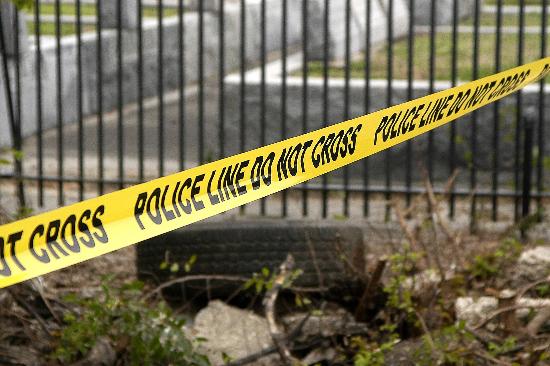 Yellow police line tape stretching diagonally across a scene with a metal fence and rubble behind it, indicating a restricted area.