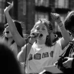 Young woman sings into a microphone with arms raised at an outdoor street performance.